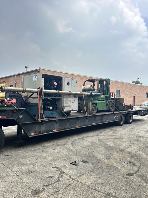 A machine and our forklift chained down together on the step deck, loaded outside a Schaumburg shop